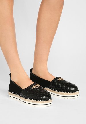 Chaussures noires en tissu matelassé à enfiler avec un accent doré. Elles présentent une tige texturée, une semelle intermédiaire en jute style espadrille et des coutures blanches contrastantes.