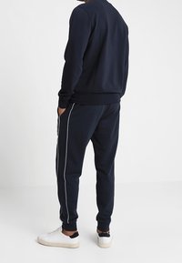 Námořnická mikina a tepláky s bílými bočními pruhy a žebrovanými manžetami. Tkanina vypadá jemně a má hladký povrch. Bílé tenisky dotvářejí vzhled.