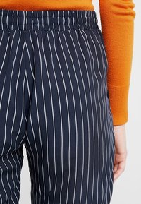 Granatowe spodnie w paski z białymi pionowymi liniami, z elastycznym paskiem w talii. Materiał jest gładki z lekkim połyskiem.