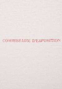 Bílá tkanina obsahuje text "COMMISSAIRE D'EXPOSITION" vytištěný středně růžovým, jednoduchým písmem bez patky ve středu.
