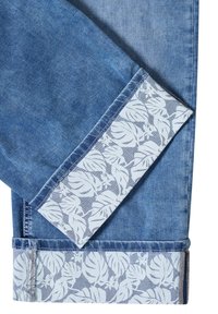 Jean en denim avec revers pliés révélant un motif de feuille tropicale blanche sur une doublure en tissu bleu clair.