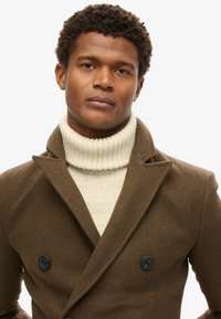 Brun dobbeltradet frakke med sorte knapper, lagt over en cremefarvet ribbet turtleneck sweater. Glat tekstur og skræddersyet design.