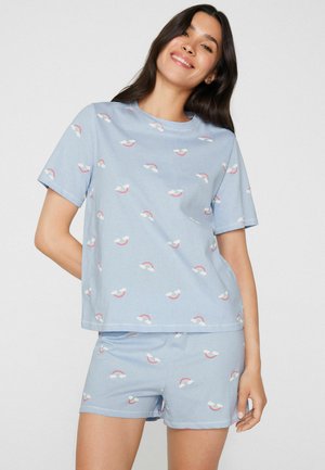 Lächelnde Frau trägt hellblaues Kurzarm-Top und Shorts mit kleinen Regenbogen- und Wolkenmustern vor einfarbigem Hintergrund.