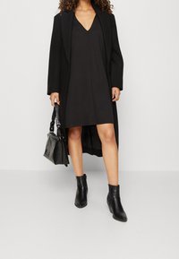 Vestido negro sin mangas con escote en V, combinado con un abrigo negro largo, botas de tobillo y un bolso negro texturizado. Tela suave, diseño minimalista.