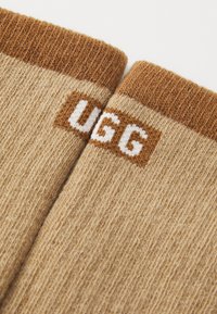 Tan färgade stickade strumpor med en brun ribbad kant och vit broderad "UGG"-logotyp på en rektangulär bakgrund. Mjuk textur, avslappnad design.