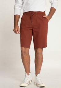 Men's brick red shorts with an elastic waistband, drawstring, side pockets, and a smooth cotton blend fabric. Worn with white high-top sneakers.