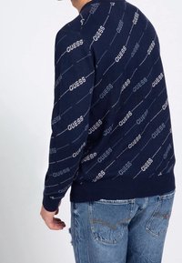 Suéter de punto azul marino con un patrón repetitivo del logotipo "GUESS", con cuello redondo y puños de canalé, combinado con jeans azules desgastados.