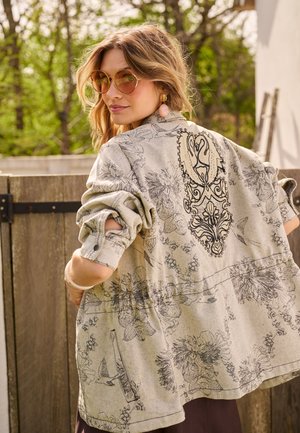 Femme portant des lunettes de soleil et une veste à motif floral avec un dessin brodé dans le dos, debout à l'extérieur près d'une clôture en bois.