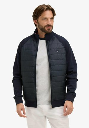 Homme portant une veste matelassée bleu marine avec fermeture éclair par-dessus une chemise gris clair, debout avec une expression neutre et regardant sur le côté.