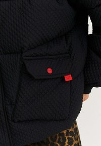 Černá strukturovaná bunda s velkou přední kapsou, která má červený patent a červenou visačku s designem řas, zkombinovaná s kalhotami s leopardím potiskem.