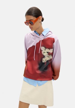 Kobieta w pomarańczowych okularach przeciwsłonecznych, fioletowo-czerwonej bluzie z kapturem w kwiaty, na niebieskiej koszuli z kołnierzykiem oraz kremowej spódnicy.