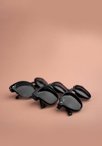 Tři páry černých slunečních brýlí zobrazené na světle růžovém pozadí. V-čkové akcenty, různé tvary zahrnují kulaté a čtvercové čočky.