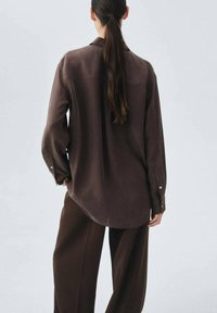 Camicia marrone a maniche lunghe con vestibilità comoda, caratterizzata da una spalla dietro, polsini con bottone e una texture liscia. Abbinata a pantaloni marroni a vestibilità larga.