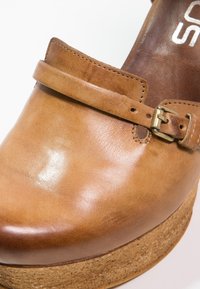 Mocassin en cuir camel avec une bride à boucle et une semelle en liège, présentant des coutures détaillées et une surface lisse et polie.