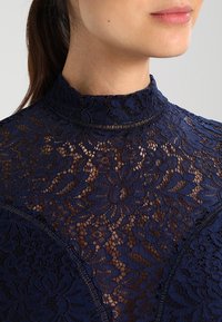 Marineblaue Spitzenbluse mit hohem Kragen, die über florale Muster und durchscheinende Textur verfügt. Sichtbare Nähte entlang der Kanten betonen das Design.