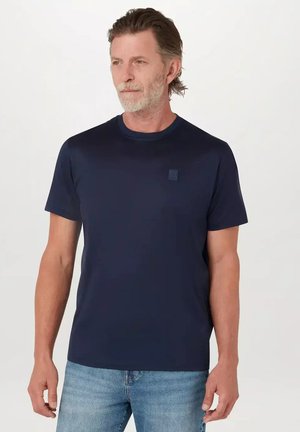 Man met grijzige baard die een marineblauw T-shirt met korte mouwen en lichtblauwe spijkerbroek draagt, staand tegen een effen witte achtergrond.