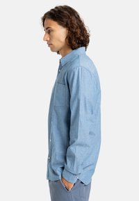Camisa de botones azul claro hecha de tela suave, con un bolsillo en el pecho izquierdo, mangas largas y un corte relajado.
