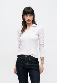 Polo a maniche lunghe di colore rosa chiaro con scollo a V e piccolo logo ricamato. Abbinata a jeans aderenti scuri. Tessuto liscio, a coste.