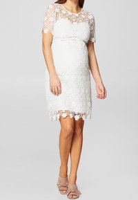 Vit spetsklänning med kort ärm, figursydd bodice och en rosett vid midjan. Vågig nederkant ger textur. Bärs med nakna sandaler.