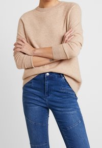 Beige gestrickter Pullover mit rundem Halsausschnitt und langen Ärmeln, kombiniert mit blauen Jeans aus Denim mit strukturiertem, tailliertem Design.