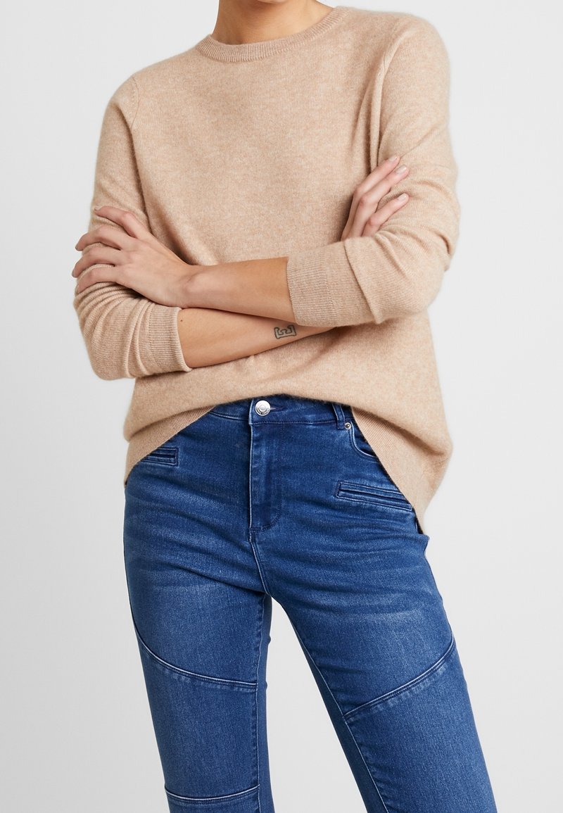 Beige gestrickter Pullover mit rundem Halsausschnitt und langen Ärmeln, kombiniert mit blauen Jeans aus Denim mit strukturiertem, tailliertem Design.