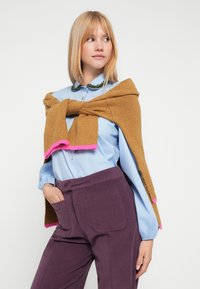 Strikket genser i sennepsgult med rosa kant, drapert over en lys blå bluse. Modellen har på seg mørke lilla bukser med sidelommer.