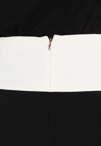Vestido en blanco y negro con un diseño estructurado, una cinturilla blanca y una cremallera central en la parte posterior con tirador plateado. Textura de tela suave.