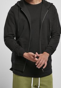 Sudadera con cremallera negra, con bolsillos frontales, puños acanalados y capucha con cordón, llevada sobre una camiseta negra y pantalones de chándal verdes.
