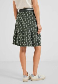 Grüner gemusterter Rock mit einem gerafften Saum, elastischem Bund und weißen Blumenmotiven. Kombiniert mit hellen Sneakers.