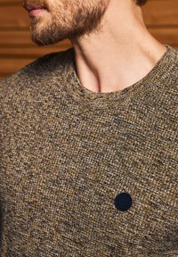 Pletený svetr v hnědých a šedých tónech, s kulatým výstřihem a texturovanou tkaninou. Obsahuje malý černý detail loga na přední straně.