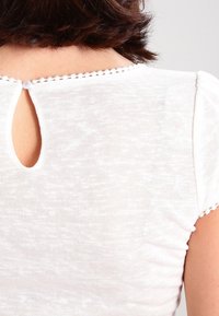 Bílý, lehký top s krátkými rukávy, klíčovou dírkou na zadní straně s knoflíkovým zapínáním a texturovanou látkou s jemnými, decentními vzory.