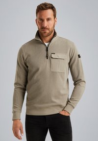 Suéter de un tono beige claro con un cuello de media cremallera, textura acanalada, un bolsillo en el pecho y detalles en negro. Las mangas y el dobladillo son ajustados.