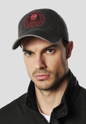 Hombre con barba incipiente que lleva una gorra negra con un emblema de calavera roja y una chaqueta negra con cremallera, mirando directamente a la cámara sobre un fondo gris.