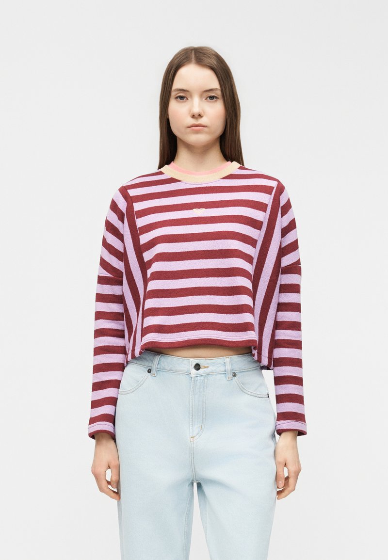 Jeune femme debout, de face, portant un pull court à manches longues rayé rose et bordeaux et un jean taille haute bleu clair.