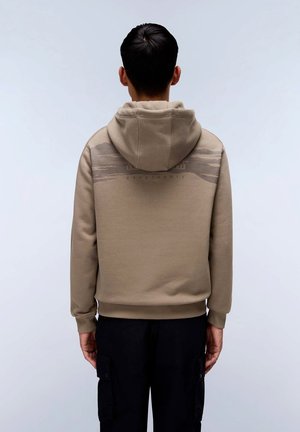 Felpa beige con cappuccio realizzata in morbido tessuto, caratterizzata da un motivo sottile sulla parte superiore della schiena e polsini a coste. Indossata sopra pantaloni scuri.