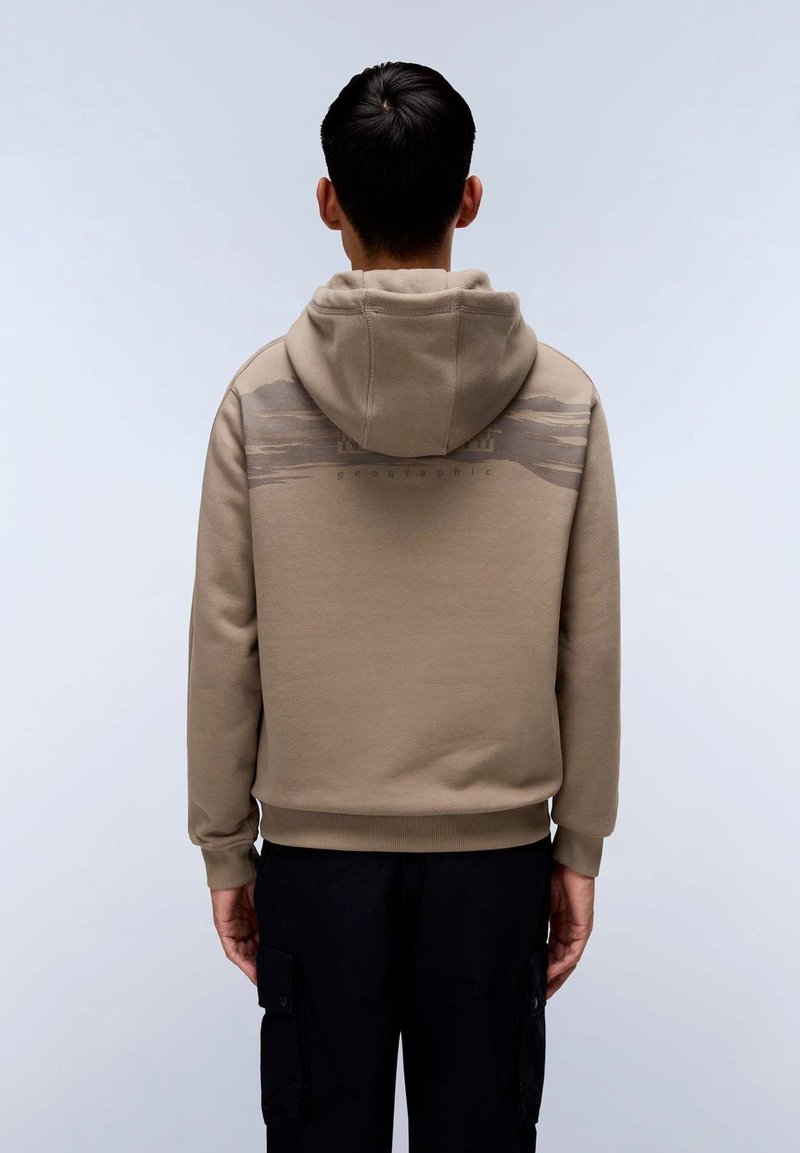 Felpa beige con cappuccio realizzata in morbido tessuto, caratterizzata da un motivo sottile sulla parte superiore della schiena e polsini a coste. Indossata sopra pantaloni scuri.