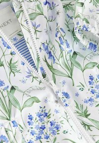 Tissu blanc avec motif floral bleu et vert, comprenant une fermeture éclair et un détail de poignet élastique sur la manche du vêtement.