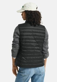 Schwarze Pufferweste mit gestepptem Design und hohem Kragen, über einem grauen, strukturierten Pullover getragen. Das Model trägt hellblaue Jeans.