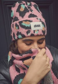 Rozā leoparda raksta adīta beanie cepure ar tirkīza, oranžiem un melniem akcentiem. Saskaņota šalle piedāvā līdzīgu rakstu, abas izgatavotas no maiga, teksturēta auduma.