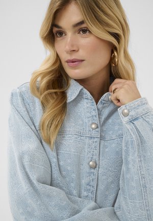 Une femme blonde aux cheveux lâches et ondulés porte une veste en denim bleu clair texturée avec des boutons semblables à des perles et des boucles d'oreilles créoles dorées.