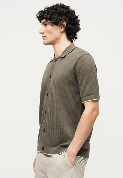 Άνδρας με σγουρά μαλλιά στέκεται πλευρικά φορώντας μια χιτωνα με κοντά μανίκια σε ελαιοπράσινο χρώμα και ανοιχτό μπεζ παντελόνι με το χέρι στην τσέπη.