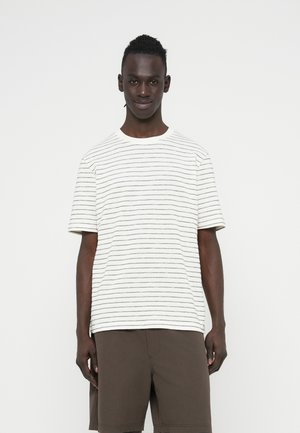 Maglietta a maniche corte a righe bianca e blu navy, con scollo rotondo. Il tessuto sembra morbido con una vestibilità comoda. Indossata con pantaloncini marroni.