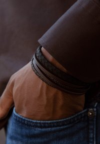 Drie leren armbanden in donkerbruin, met ingeweven en gladde texturen, gedragen om een pols die gedeeltelijk zichtbaar is in een denim zak.