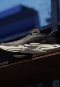 Schwarzer Sportschuh mit weißer, gepolsterter Sohle, der auf einer Holzoberfläche vor einem verschwommenen blauen und dunklen Hintergrund liegt.