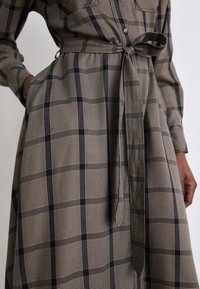 Femme portant une robe à carreaux marron et noir avec une ceinture nouée à la taille et une main dans une poche.