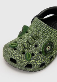 Roheline crocs-stiilis king, millel on praguline tekstuur, rohelised aktsendid, tõstetud elemendid ja silmad külgedel. Avatud disain ventilatsiooniavadega.