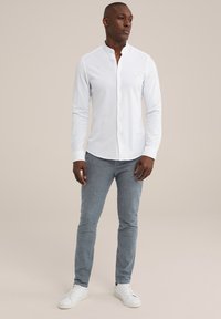 Camisa blanca de manga larga con cuello de banda, con botones al frente, combinada con pantalones gris ajustados y zapatillas blancas. Diseño limpio y minimalista.