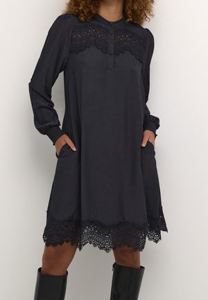 Zwarte jurk met lange mouwen, afgewerkt met kant bij de borst en zoom, knoopsluiting aan de voorkant, zakken, gedragen met zwarte kniehoge laarzen.