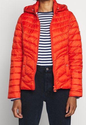 Vrouw draagt een feloranje gewatteerde jas over een marineblauw-wit gestreept shirt en donkere broek, staat met handen ontspannen langs het lichaam.