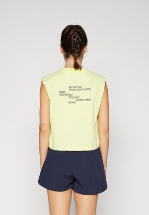 Kobieta z warkoczami, ubrana w bladożółty top bez rękawów z motywacyjnym napisem oraz granatowe krótkie spodenki, stoi tyłem na jednolitym tle.